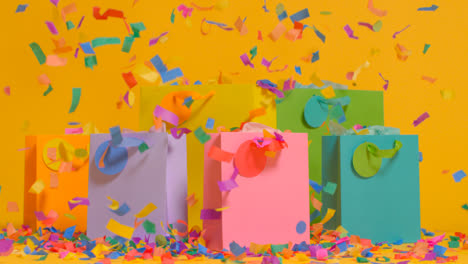 Studio-Shot-Of-Colourful-Birthday-Party-Gift-Bags-Against-Yellow-Background-With-Falling-Paper-Confetti-1